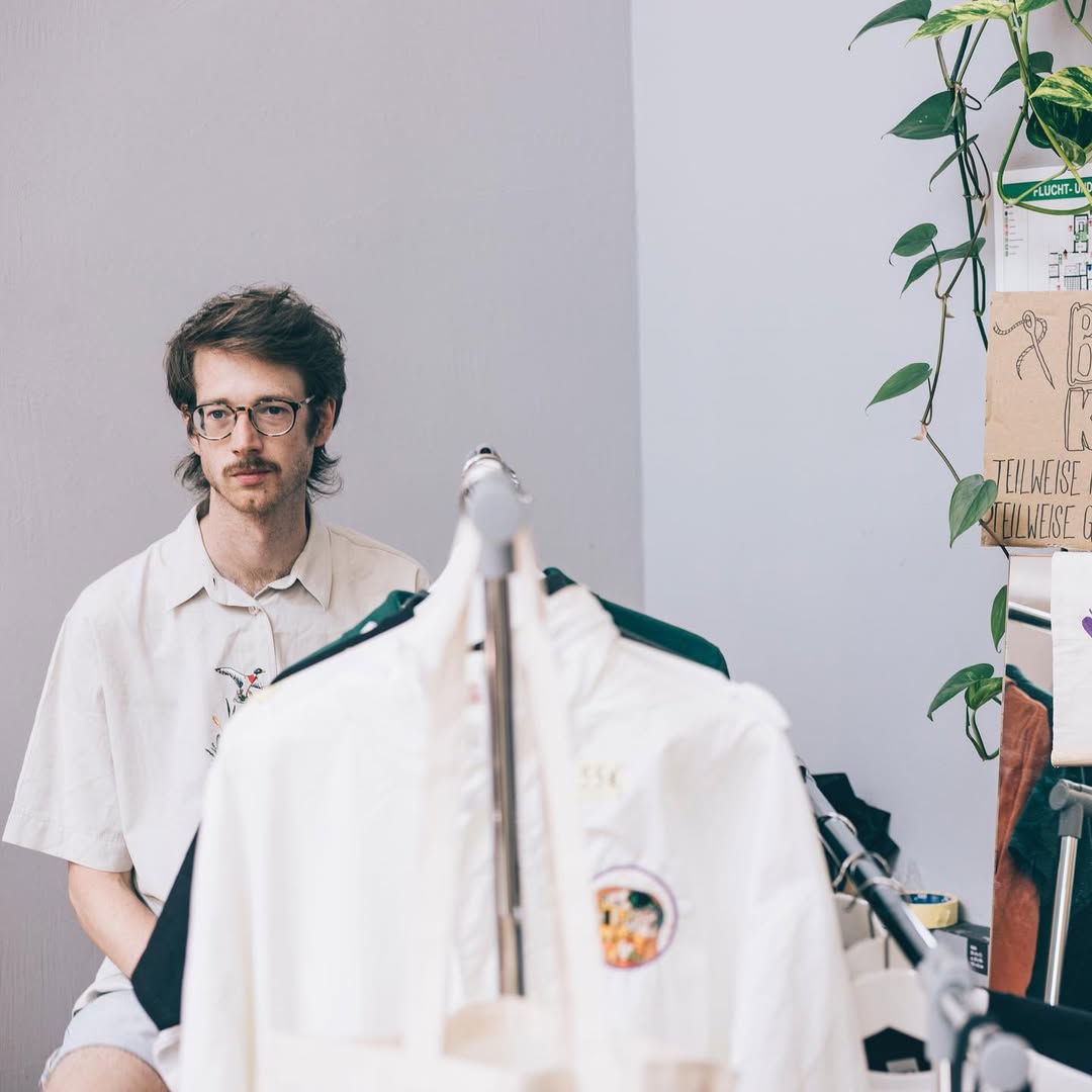 stickolas Nikolas Becker sitzt im hinteren Bereich des Bildes und schaut links neben der Kamera vorbei. Im Vordergrund unscharf steht ein Kleiderständer mit Hemden in weiß und grün. Bild: Sarah Hähnle
