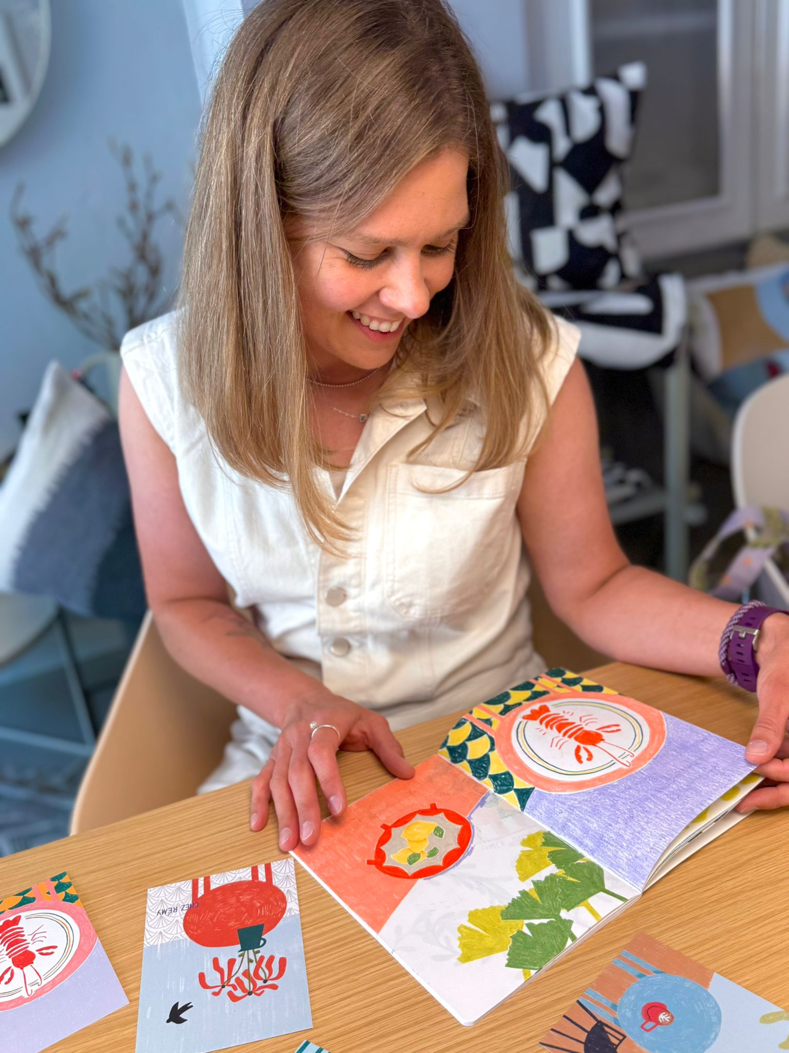 Eva_Czajkowsky_by_Alexandra_ebert-roedel Eva Czajkowski sitzt lächelnd an einem Holztisch. Vor ihr liegen Postkarten und ein Buch mit bunten Bildern (Bild: Alexandra Ebert-Roedel).
