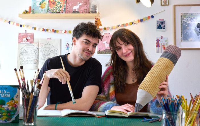 Sam Rassy (links) und Juliana Kralik (rechts) sitzen an einem Tisch und lächeln in die Kamera. Sam hält einen langen Pinsel in der rechten, Juliana einen großen Stift aus Plüsch in der linken Hand (Bild von Neri Kranz)
