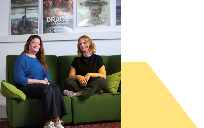 Nicola Dahlke (links) und Julia Knopp (rechts) sitzen auf einer grünen Couch und lächeln in die Kamera. An der Wand im Hintergrund hängen gerahmte Plakate von Dokumentationen und filmen (Bild von Neri Kranz).